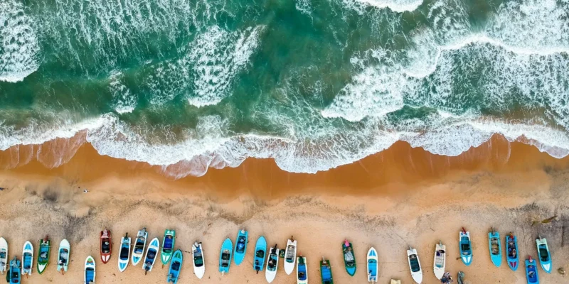 An aerial top-down view of a long row of colorful fishing boats parked on the golden sand of Arugam Bay next to crashing turquoise waves in Sri Lanka