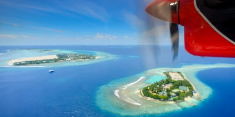 View from a seaplane window showing the red propeller engine flying over tropical coral islands and turquoise lagoons in the Maldives