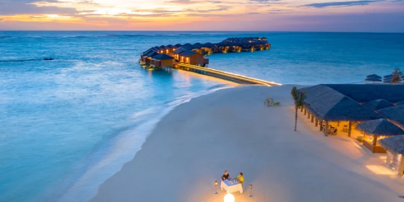 Aerial view of a couple enjoying a private candlelit beach dinner at sunset next to an overwater villa resort in the Maldives