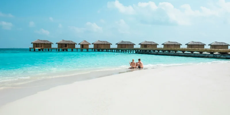 A couple sitting on a pristine white sand beach next to a turquoise ocean with overwater bungalows in the Maldives