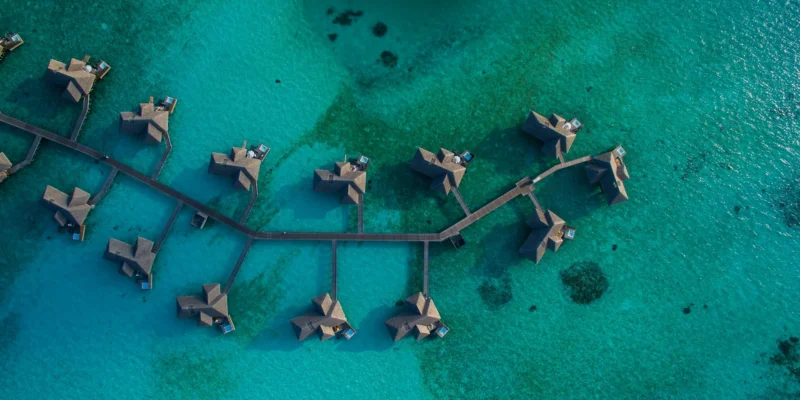 Top-down aerial view of luxury overwater villas branching off a wooden jetty in a clear turquoise lagoon in the Maldives